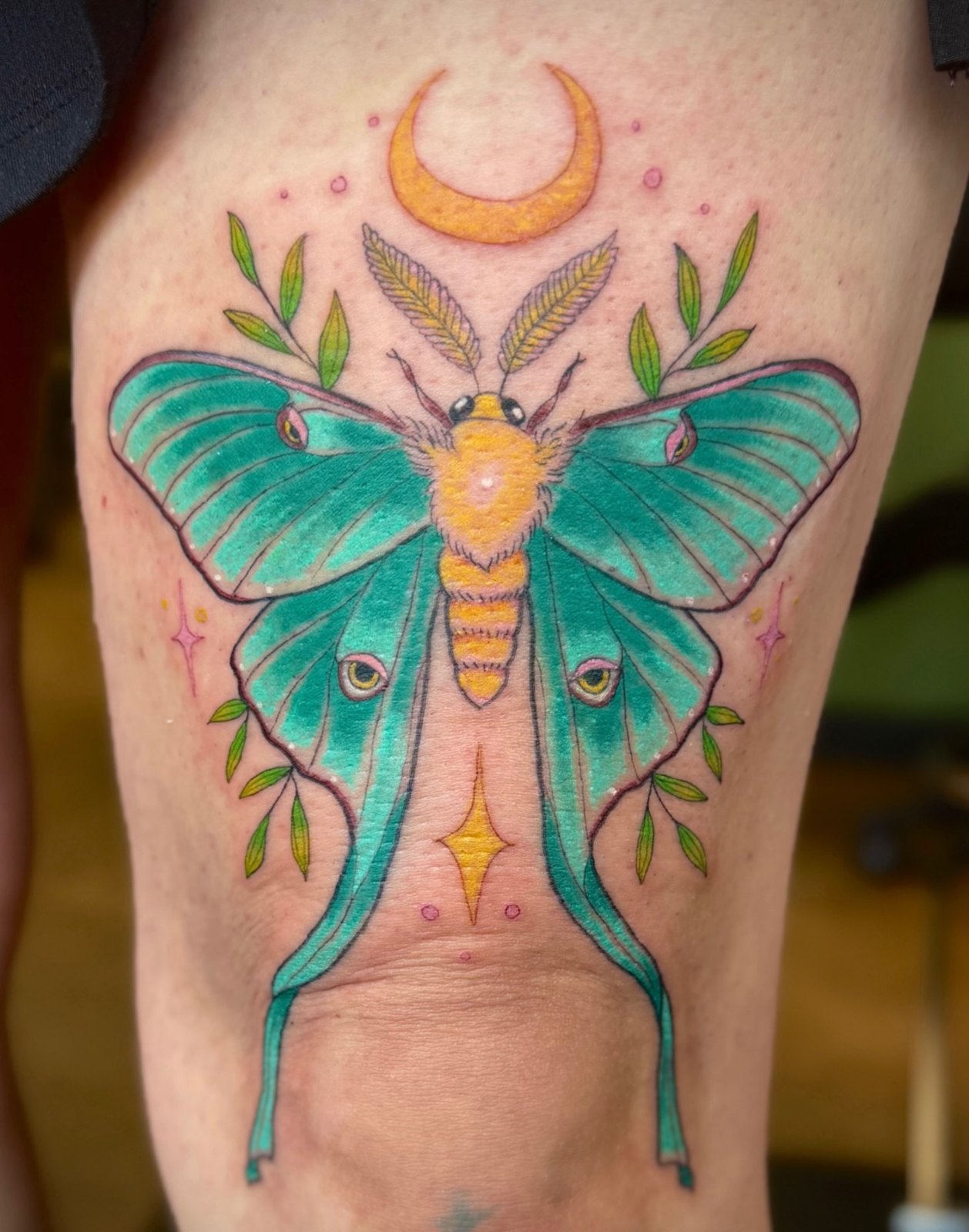 Colour tattoo of a teal luna moth with a gold crescent moon and green sprigs